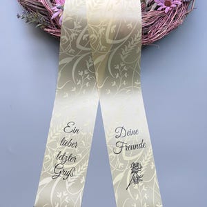 Könnte beinhalten: Cremefarbenes Band mit floralem Muster und schwarzer Fransenborte. Auf dem Band steht "Ein lieber letzter Gruß" und "Deine Freunde" mit einer Rosenillustration. Das Band liegt über einem Kranz mit rosa und lila Blüten.