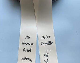 Puede incluir: Cinta de raso marfil con texto y flecos negros. El lado izquierdo dice "Als letzten Gruß" con una ilustración de ramita. El lado derecho dice "Deine Familie" con una ilustración de diente de león. Una corona floral está en el fondo.