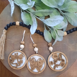 May include: Three circular wooden ornaments featuring laser-cut designs of a dragonfly, a butterfly, and a hummingbird with flowers. Each ornament has a white resin inlay, a wooden bead, and a white string. A black wooden bead garland is included.