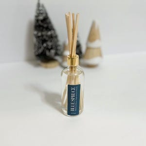 May include: A clear glass diffuser bottle with a gold cap and wooden reeds. The label reads "BLUE SPRUCE". The bottle is filled with a clear liquid. Decorative Christmas trees are in the background.