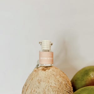 May include: A small glass bottle with a wooden cap and a pink label that says "Island Mango" on it. The bottle is sitting on a coconut and a mango.