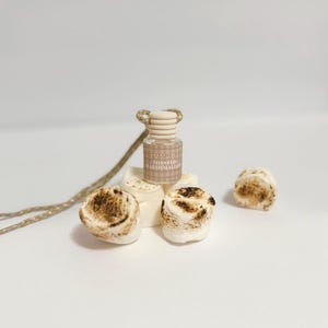 May include: A small glass bottle with a ribbed wooden cap and a beige gingham label reading Toasted Marshmallow, surrounded by several white marshmallows with browned, toasted tops, attached to a thin, light-colored braided hanging cord.
