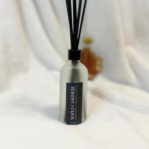 May include: A frosted glass diffuser bottle with black reeds. The label reads "MAPLE CASHMERE". The bottle has a black cap and is filled with a liquid. The background is a soft, white fabric.
