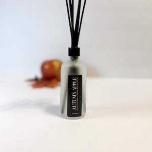 May include: A frosted glass diffuser bottle with black reeds. The label reads "AUTUMN APPLE" in white text. An apple and fall leaves are in the background. The diffuser is handcrafted in Nebraska.