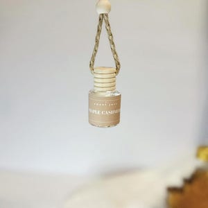 May include: A small, clear glass bottle with a wooden cap and a beige label that reads "MAPLE CASHMERE." The bottle is suspended by a braided rope and a wooden bead. The product is likely an air freshener or fragrance.