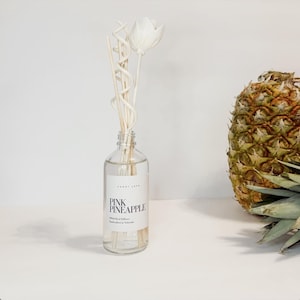 May include: A clear glass bottle with a white label that reads "PINK PINEAPPLE" and "Reed Diffuser" is filled with a clear liquid and has white wooden sticks with a white flower on top. A pineapple is in the background.