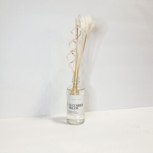 May include: A clear glass bottle diffuser with a white label that reads "Cucumber Melon" and natural reed sticks. The sticks are topped with white dried flowers.