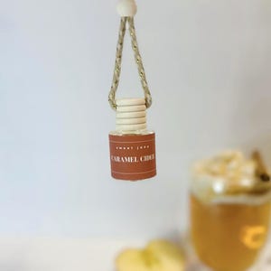 May include: A small, clear glass bottle with a wooden cap and a brown label that reads "sweet jane CARAMEL CIDER." The bottle is suspended by a rope with gold accents. The background is white, with a blurred image of a drink.