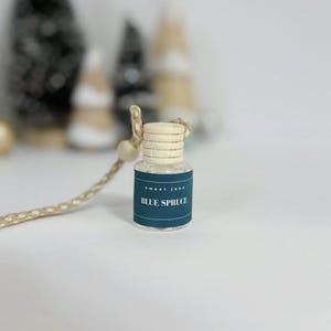 May include: A small, clear glass bottle with a wooden cap and a blue label that reads "BLUE SPRUCE." The bottle is suspended by a gold-colored string with a wooden bead. The background is blurred, with hints of Christmas tree decorations.