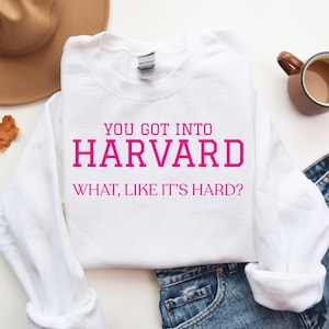 May include: A white sweatshirt with the text "YOU GOT INTO HARVARD WHAT, LIKE IT'S HARD?" in pink. The sweatshirt is paired with blue jeans, a brown hat, and a mug of coffee. An autumn leaf is also present.