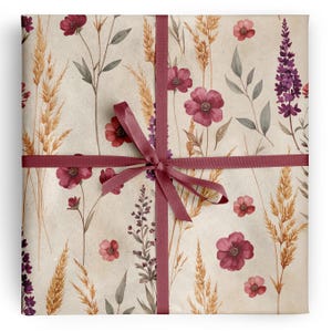 May include: A square gift package wrapped in light beige paper featuring a botanical print of dark pink flowers, purple spikes, and golden wheat stalks. A dark pink satin ribbon is tied in a cross pattern around the center of the package.