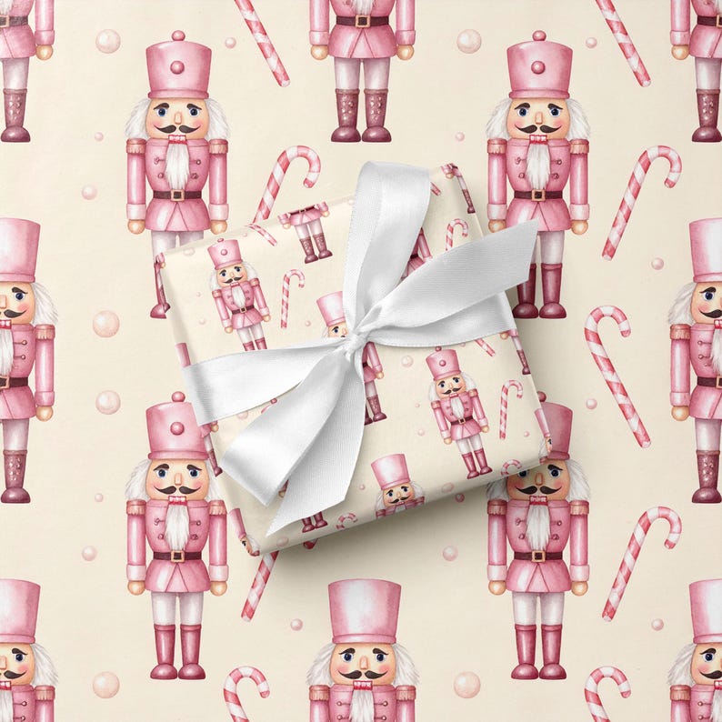 May include: A rectangular gift box wrapped in cream-colored paper featuring a repeating pattern of pink nutcrackers and red and white candy canes, tied with a wide, white satin ribbon bow centered on the top surface of the package.