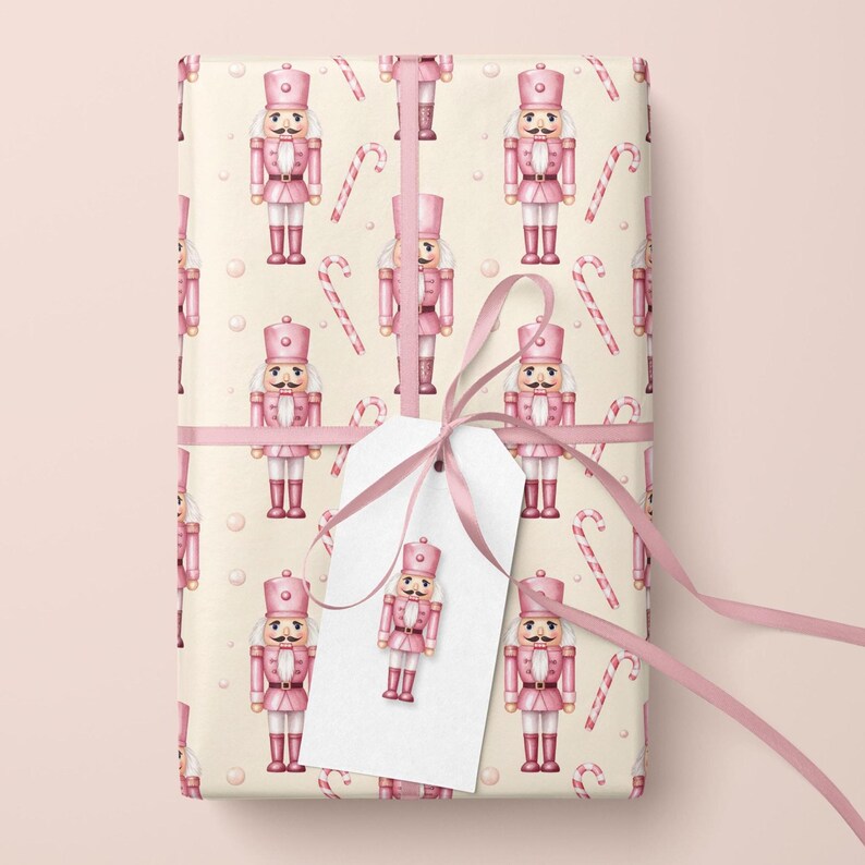 May include: A rectangular gift wrapped in cream-colored paper featuring a repeating pattern of pink nutcrackers and red and white candy canes. A pink satin ribbon is tied around the package, securing a blank white gift tag with a nutcracker.