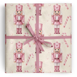 May include: A square gift package wrapped in light beige paper featuring a repeating pattern of pink nutcracker figures and red and white candy canes, tied with a pink satin ribbon centered in a bow across the middle of the package.