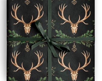 May include: A square gift package wrapped in black paper featuring a repeating pattern of brown deer heads with large antlers, green pine branches, and gold decorative flourishes, tied with a dark green satin ribbon in a centered bow.