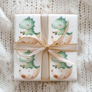 May include: A wrapped gift with a white background featuring a repeating pattern of green baby dinosaurs hatching from eggs. The gift is tied with a gold ribbon and bow, perfect for a baby shower or birthday.