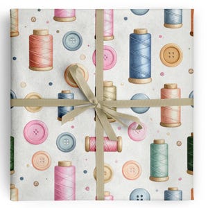May include: A wrapped gift featuring a repeating pattern of watercolour spools of thread and buttons in shades of pink, blue, green, and tan on a white background. The gift is tied with a beige ribbon.