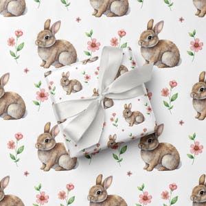 May include: White gift wrap featuring a repeating pattern of brown bunnies and pink flowers. A wrapped gift with a white satin bow is in the foreground. The overall design is a charming, spring-themed pattern.