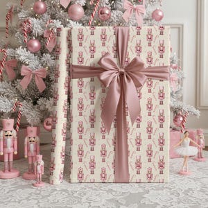 May include: A rectangular gift wrapped in cream-colored paper featuring a repeating pattern of pink nutcrackers and candy canes, tied with a wide, dusty pink satin ribbon bow. The gift is positioned upright against a white Christmas tree.