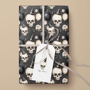 May include: Wrapped gift with a black and white skull pattern, party hats, balloons, and roses. A white tag with a skull illustration and the text "Fifty + Fabulous" is attached with a gray ribbon. The wrapping paper has a dark background.