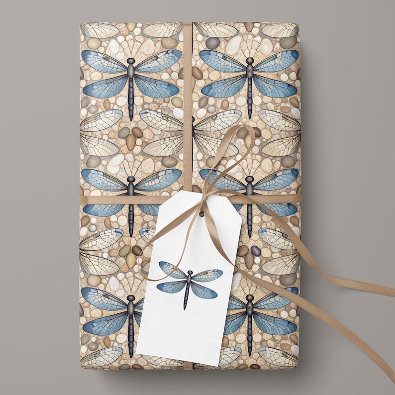 May include: Gift-wrapped package featuring a repeating pattern of blue dragonflies and pebbles on a beige background. The package is tied with a brown ribbon and a white tag with a dragonfly illustration.
