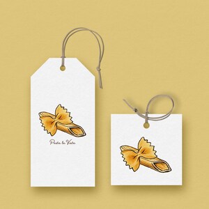 Pode incluir: Duas etiquetas de presente brancas com uma ilustração em aquarela de massa farfalle. A etiqueta maior tem o texto "Pasta la Vista". Ambas as etiquetas têm um fio marrom preso para pendurar, em um fundo amarelo.