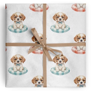 Puede incluir: Papel de regalo blanco con un patrón repetido de cachorros de acuarela con pajaritas azules, sentados en flotadores a cuadros. El papel está atado con una cinta y un lazo marrón.