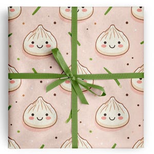 May include: Gift wrap featuring a repeating pattern of smiling dumpling illustrations on a light pink background. The dumplings have a white exterior and a pink blush. Green ribbon is tied around the gift wrap in a bow.