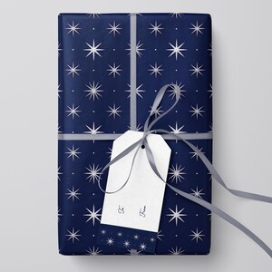 Peut inclure: Un cadeau emball&eacute; avec du papier bleu marine orn&eacute; d'un motif d'&eacute;clats d'&eacute;toiles blancs. Une &eacute;tiquette cadeau blanche est attach&eacute;e avec un ruban gris. L'&eacute;tiquette porte les mots "&Agrave;" et "De".