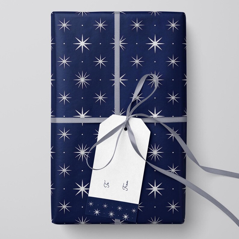 Peut inclure: Un cadeau emball&eacute; avec du papier bleu marine orn&eacute; d'un motif d'&eacute;clats d'&eacute;toiles blancs. Une &eacute;tiquette cadeau blanche est attach&eacute;e avec un ruban gris. L'&eacute;tiquette porte les mots "&Agrave;" et "De".