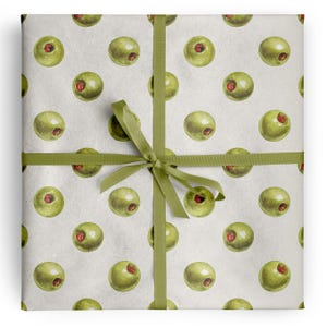 Könnte beinhalten: Ein quadratisches Geschenkpaket, eingewickelt in cremefarbenes Papier mit einem sich wiederholenden Muster aus grünen Oliven mit roten Pimentostücken. Das Paket ist mit einem olivgrünen Satinband umwickelt, das eine mittige Schleife bildet.
