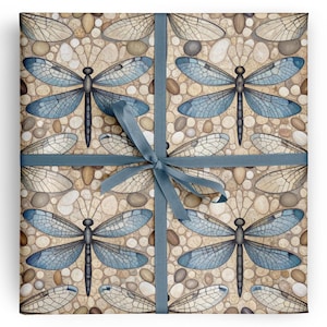 May include: Square gift wrap featuring a repeating pattern of blue dragonflies and dragonfly wings on a pebble background. A blue ribbon is tied around the wrapped package. The dragonfly wings are detailed with delicate veins.