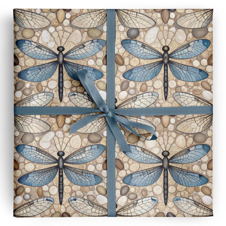 May include: Square gift wrap featuring a repeating pattern of blue dragonflies and dragonfly wings on a pebble background. A blue ribbon is tied around the wrapped package. The dragonfly wings are detailed with delicate veins.
