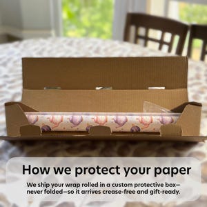 May include: A roll of wrapping paper featuring a repeating pattern of pastel-colored teapots and small stars, resting inside an open brown cardboard shipping box. The text on the image reads: How we protect your paper. We ship your wrap rolled in a custom protective box&mdash;never folded&mdash;so it arrives crease-free and gift-ready.