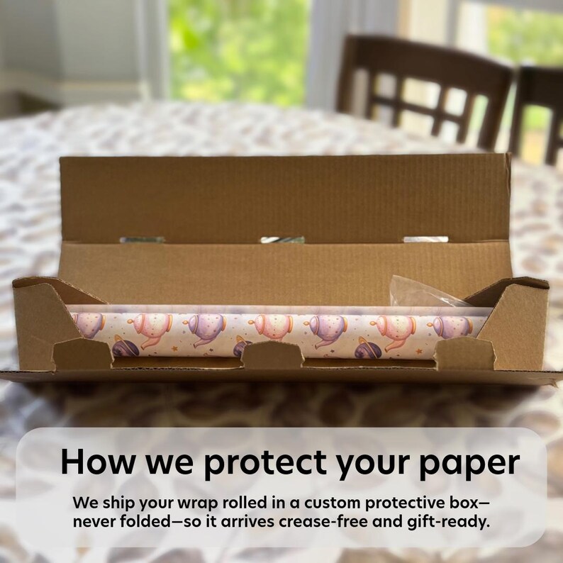 May include: A roll of wrapping paper featuring a repeating pattern of pastel-colored teapots and small stars, resting inside an open brown cardboard shipping box. The text on the image reads: How we protect your paper. We ship your wrap rolled in a custom protective box&mdash;never folded&mdash;so it arrives crease-free and gift-ready.