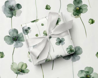 Può includere: Un regalo confezionato con un fiocco di nastro di raso bianco. La carta da regalo presenta un motivo floreale ad acquerello di fiori verdi e grigi su sfondo bianco. I fiori hanno centri verde scuro e lunghi steli verdi.