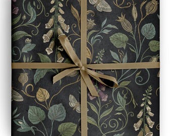 Op de afbeelding: Een vierkant pakket verpakt in donkergrijs papier met een botanisch patroon van gedempte groene bladeren, crèmekleurige vingerhoedskruidbloemen en motten. Het pakket is vastgebonden met een dun, glad, lichtbruin lint in een kruisvorm.