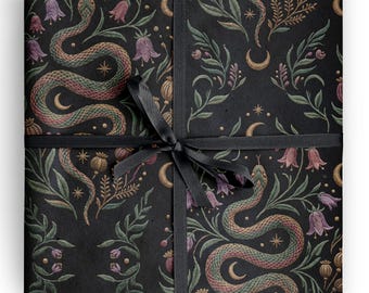 Op de afbeelding: Een vierkant cadeaupakket verpakt in zwart papier met een herhalend patroon van kronkelende slangen, paarse klokvormige bloemen, groen gebladerte, gouden maansikkels en sterren, vastgebonden met een dun zwart satijnen lint in een strik.