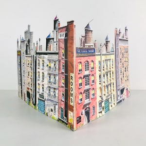 May include: A three-dimensional paper model of a city block featuring multiple buildings with brick and stone facades. The structure displays text including HOTEL, ROOMS, and GENERAL STORE on various building fronts with fire escapes.