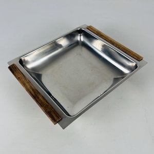 May include: A square stainless steel serving tray with a polished silver finish and two parallel wooden handles attached to opposite sides. The tray features a deep, recessed center and a flat, wide rim that extends to the wooden accents.