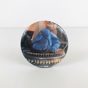 May include: A blue alien creature with a long nose sits on a round, metallic platform. The creature has large, round eyes and a small mouth. The platform has a series of small, round holes around the edge.