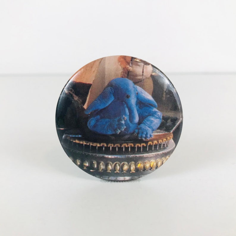 May include: A blue alien creature with a long nose sits on a round, metallic platform. The creature has large, round eyes and a small mouth. The platform has a series of small, round holes around the edge.
