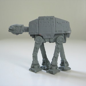 May include: A gray plastic toy model of an AT-AT walker from the Star Wars franchise. The model has four legs and a large body with a detailed design.