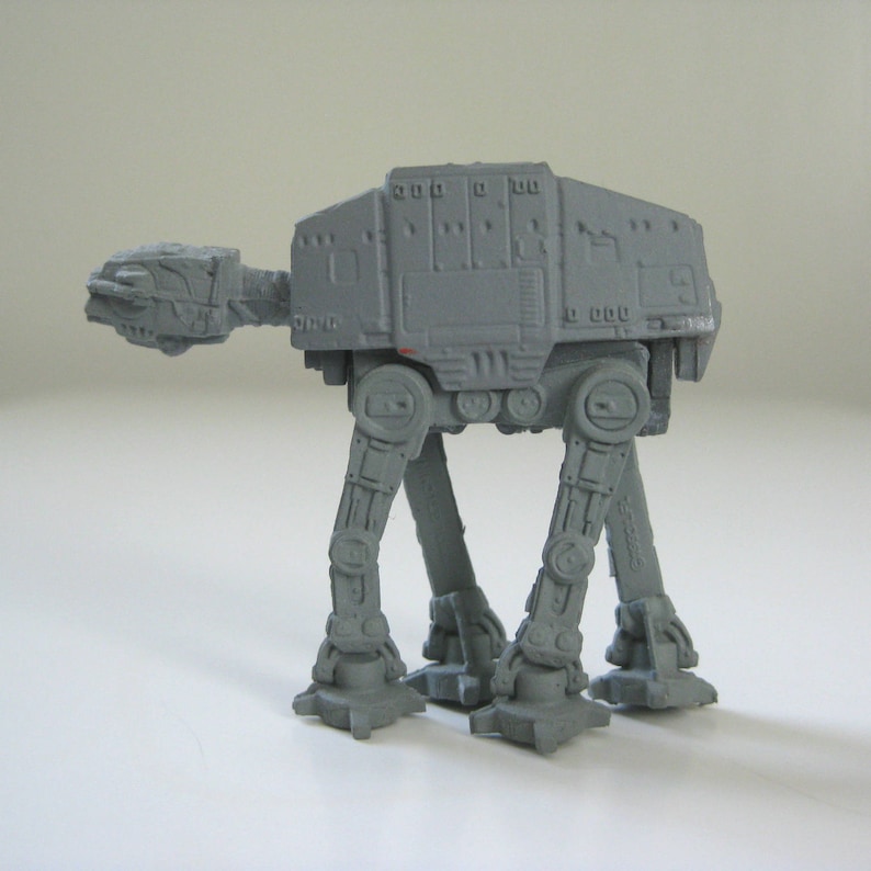 May include: A gray plastic toy model of an AT-AT walker from the Star Wars franchise. The model has four legs and a large body with a detailed design.