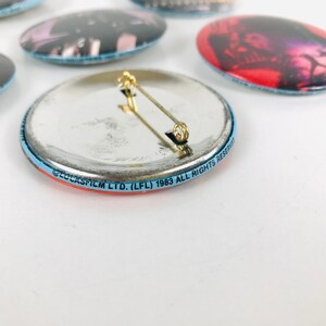 May include: The back of a round, blue and red button with a silver back. The button has a pin on the back and the text "LUCASFILM LTD. (LFL) 1983 ALL RIGHTS RESERVED" printed around the edge.