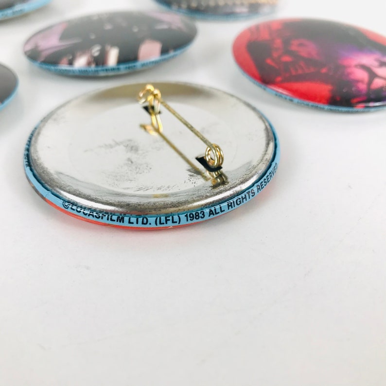 May include: The back of a round, blue and red button with a silver back. The button has a pin on the back and the text "LUCASFILM LTD. (LFL) 1983 ALL RIGHTS RESERVED" printed around the edge.