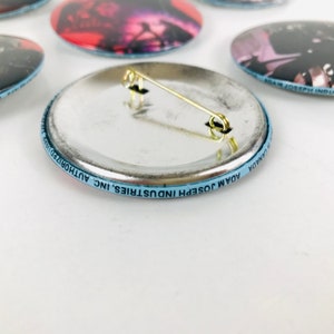 May include: The back of a round, silver button with a blue border. The border has the text "ADAM JOSEPH INDUSTRIES INC. AUTHORIZED" and "ADAM JOSEPH INDUSTRIES INC. AUTHORIZED" printed on it. The button has a safety pin attached to it.
