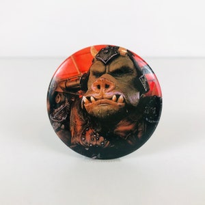 May include: A round button featuring a close-up of a brown and green creature with a large snout and sharp teeth. The creature is wearing a dark brown and black outfit with a red and orange background.
