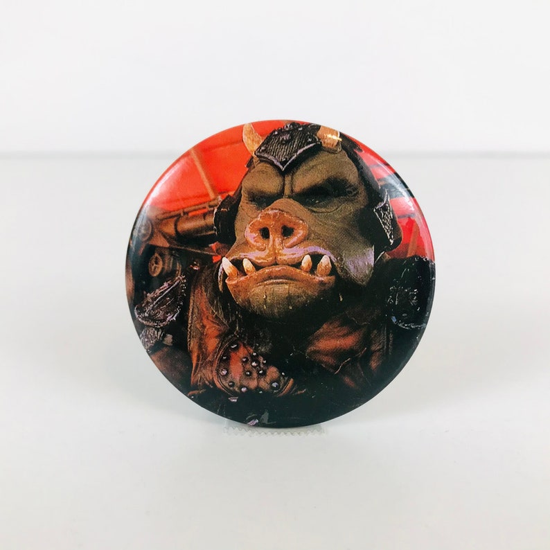 May include: A round button featuring a close-up of a brown and green creature with a large snout and sharp teeth. The creature is wearing a dark brown and black outfit with a red and orange background.