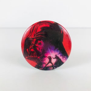 May include: A red, white, and black button featuring a silhouette of Darth Vader from Star Wars. The button shows a scene of Darth Vader and Luke Skywalker fighting with lightsabers.